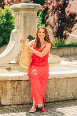 Summer portrait of happy woman with beautiful hair style, wearing red maxi dress, resting by the fountain