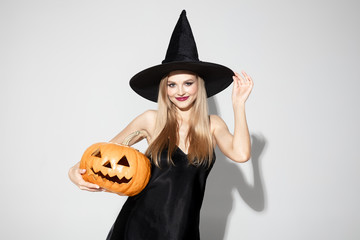 Young blonde woman in black hat and costume on white background. Attractive caucasian female model posing. Halloween, black friday, cyber monday, sales, autumn concept. Copyspace. Holding a pumpkin.