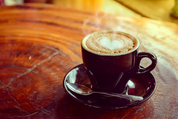 Hot coffee, heart shape with milk latte on top of black ceramic cup with metal spoon on wooden table in coffee shop in vintage tone, copy space for your text. Times to relax with caffiene concept.