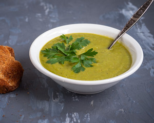 Pea soup mashed in a deep white plate with fresh sprigs of parsley, spoon and fried toast on a gray background