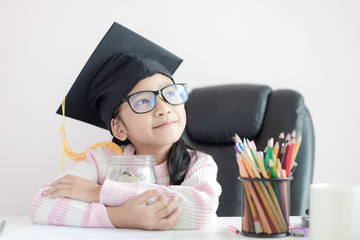 Little Asian girl wearing graduate hat hugging clear glass jar piggy bank and smile with happiness for money saving to wealthness succesful in the future of education concept select focus