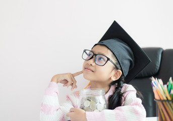 Little Asian girl wearing graduate hat hugging clear glass jar piggy bank and smile with happiness for money saving to wealthness succesful in the future of education concept select focus