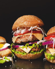 Concept: fast food big burgers on a black table. Large double cheeseburger with beef cutlets and fresh vegetables, composition: pepper, onion, tomatoes,egg, salad, cheese on a dark background.