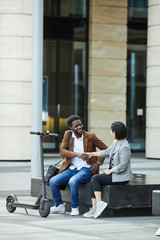 Portrait of smiling African-American man shaking hands with Asian woman while sitting on bench outdoors, copy space