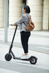 Full length side view of young Asian woman riding electric scooter in city street with architecture in background, copy space