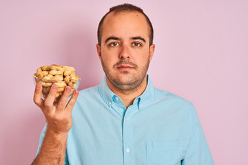 Young man holding bowl with peanuts standing over isolated pink background with a confident expression on smart face thinking serious