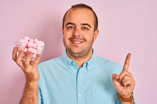 Young man holding bowl with marshmallows standing over isolated pink background surprised with an idea or question pointing finger with happy face, number one