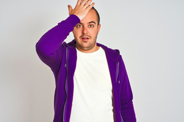 Young man wearing purple sweatshirt standing over isolated white background surprised with hand on...