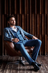 Young successful businessman in a blue suit in a cage is sitting in a leather chair. young man with a beard smiling and looks into the distance