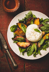 Flat-lay of seasonal salad with Burrata soft cheese and grilled peaches with glass of rose wine over rustic red painted table background, top view, close-up