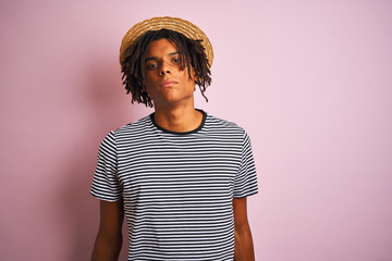 Afro american man with dreadlocks wearing navy t-shirt and hat over isolated pink background with serious expression on face. Simple and natural looking at the camera.