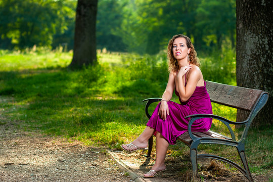 Beautiful curly hair young woman wearing elegant purple dress sitting on a bench in the middle of forest with rays of sunlight during sunset waiting her love
