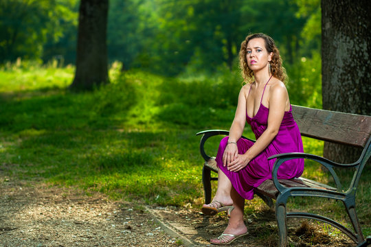 Beautiful curly hair young woman wearing elegant purple dress sitting on a bench in the middle of forest with rays of sunlight during sunset waiting her love