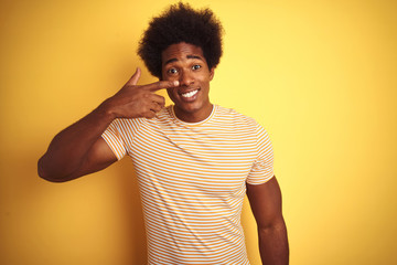 American man with afro hair wearing striped t-shirt standing over isolated yellow background Pointing with hand finger to face and nose, smiling cheerful. Beauty concept