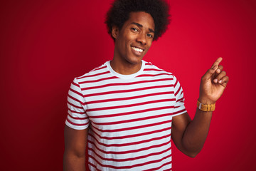 Fototapeta premium Young african american man with afro hair wearing striped t-shirt over isolated red background with a big smile on face, pointing with hand and finger to the side looking at the camera.