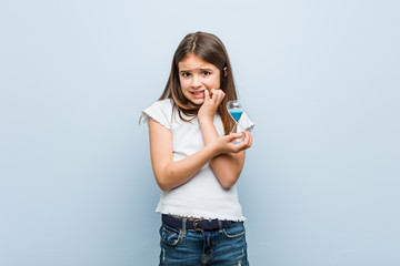 Little caucasian girl holding an hourglass biting fingernails, nervous and very anxious.