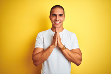 Young caucasian man wearing casual white t-shirt over yellow isolated background praying with hands together asking for forgiveness smiling confident.