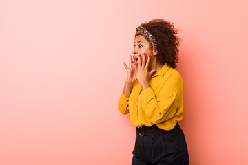 Young african american woman against a pink background shouts loud, keeps eyes opened and hands tense.