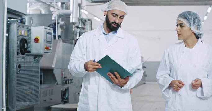 In a big manufacturing industry two young engineers walking through the industrial machine and analyzing the plan of security they looking through the map and chatting with each other