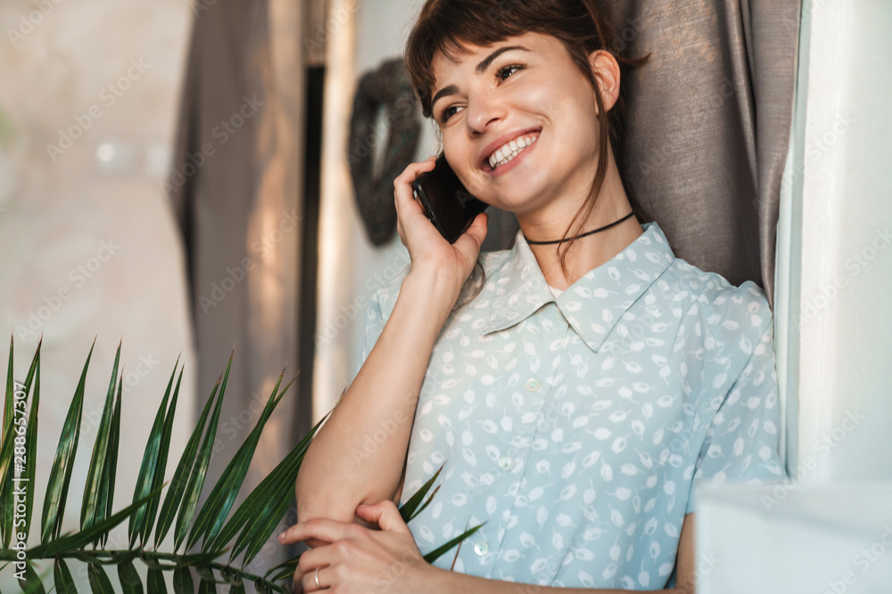 Canvas Prints Cheerful pleased happy young beautiful woman talking by mobile phone indoors at home.