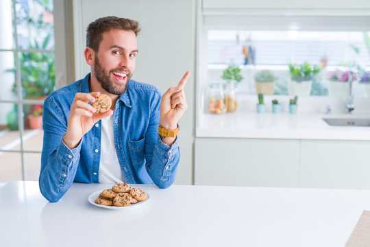 Handsome man eating chocolate chips cookies very happy pointing with hand and finger to the side