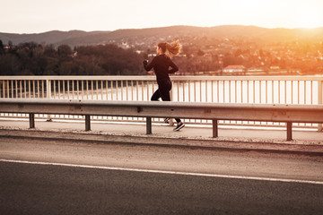 Young woman in black sports outfit running on the bridge in the city during sunset.