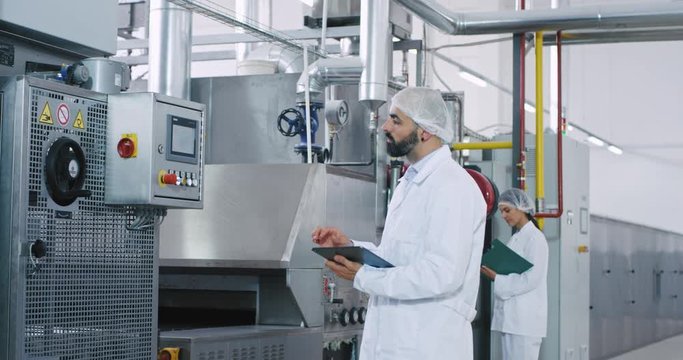 In a big manufacturing industry two engineers with the maps and electronic tablet making some notes while checking the industrial machine