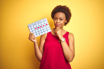 Young african american woman holding menstruation calendar over isolated yellow background serious face thinking about question, very confused idea