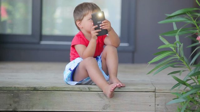 Little cute boy sits on the porch of the house and takes a pictures with mobile phone