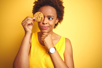 African american woman holding chocolate chips cookie over yellow background serious face thinking about question, very confused idea