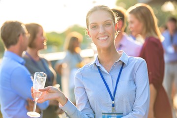 Portrait of young smiling businesswoman standing while holding wine glass against colleagues at success party on rooftop
