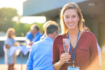 Portrait of young smiling businesswoman standing while holding wine glass against colleagues at success party on rooftop