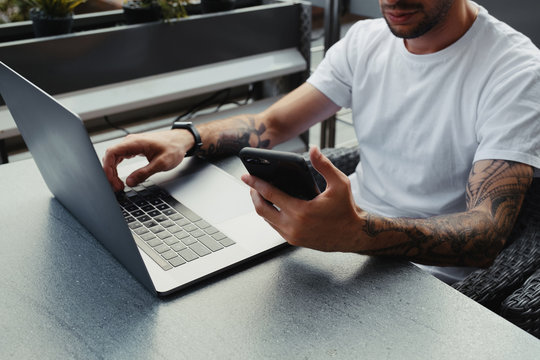 Handsome bearded guy with tattooed hands dressed white shirt using smartphone and notebook computer in outdoors coffee shop, male holding cell phone and typing text on laptop keyboard.