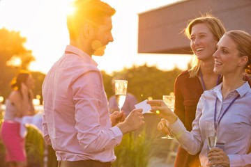 Young businesswoman giving business card to colleague while holding wine glass on success party at rooftop