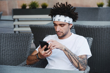 Handsome man holding modern smartphone dialing number, male with dreadlocks hair in white t-shirt using mobile phone for chatting in social networks via 4G internet connection or wifi in outdoor cafe