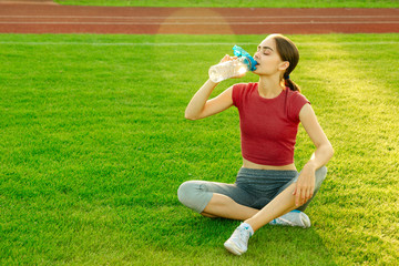 Female athlete sitting on green grass and drinking water from bottle while resting after training in stadium