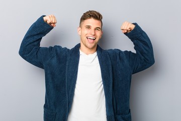 Young caucasian man wearing pajama showing strength gesture with arms, symbol of feminine power