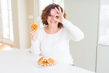 Senior woman eating sweet belgian waffle with happy face smiling doing ok sign with hand on eye looking through fingers