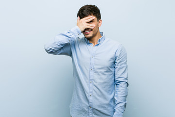 Young hispanic business man blink at the camera through fingers, embarrassed covering face.