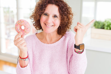 Senior woman eating pink sugar donut very happy pointing with hand and finger to the side