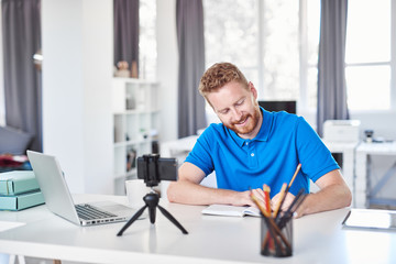 Fototapeta premium Cheerful Caucasain ginger employee sitting in his office and writing his tasks in notebook.