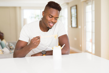 Handsome african man eating asian noodles in a delivery box, smiling enjoying lunch using chopsticks