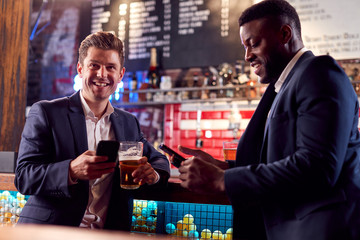 Two Businessmen Checking Mobile Phones Whilst Meeting For Drink In Bar