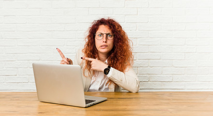 Young redhead curly woman working with her laptop shocked pointing with index fingers to a copy space.