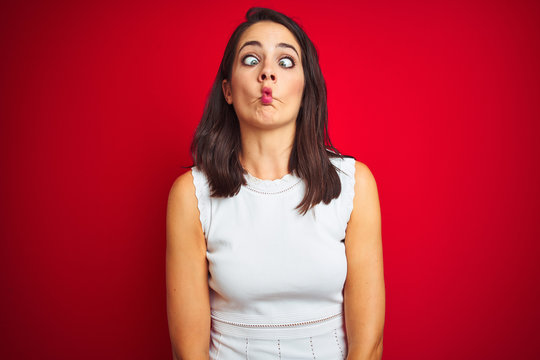 Young beautiful woman wearing white dress standing over red isolated background making fish face with lips, crazy and comical gesture. Funny expression.