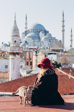 Blond woman stroking a cat on the roof with a view to the mosque, Istanbul, Turkey. Girl in a hat sits on the roof in Istanbul, sunny autumn day. Traveler girl walks through winter Istanbul.