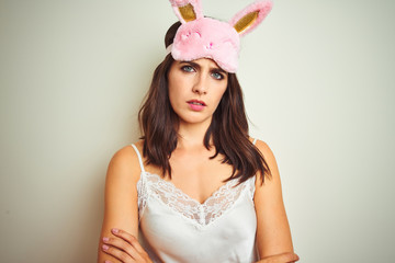 Young beautiful woman wearing pajama and mask standing over white isolated background skeptic and nervous, disapproving expression on face with crossed arms. Negative person.