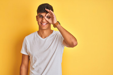 Fototapeta premium Young indian man wearing white t-shirt standing over isolated yellow background doing ok gesture with hand smiling, eye looking through fingers with happy face.