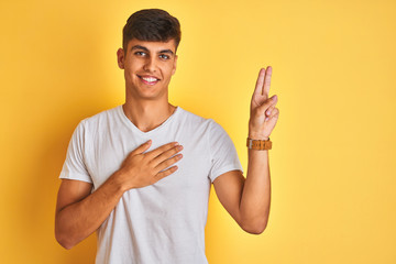 Young indian man wearing white t-shirt standing over isolated yellow background smiling swearing with hand on chest and fingers up, making a loyalty promise oath