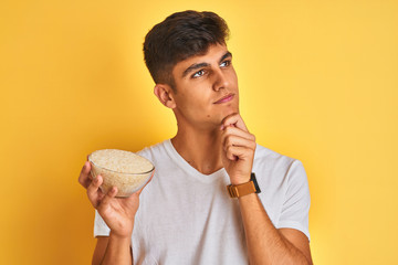 Young indian man holding bowl with rice standing over isolated yellow background serious face...